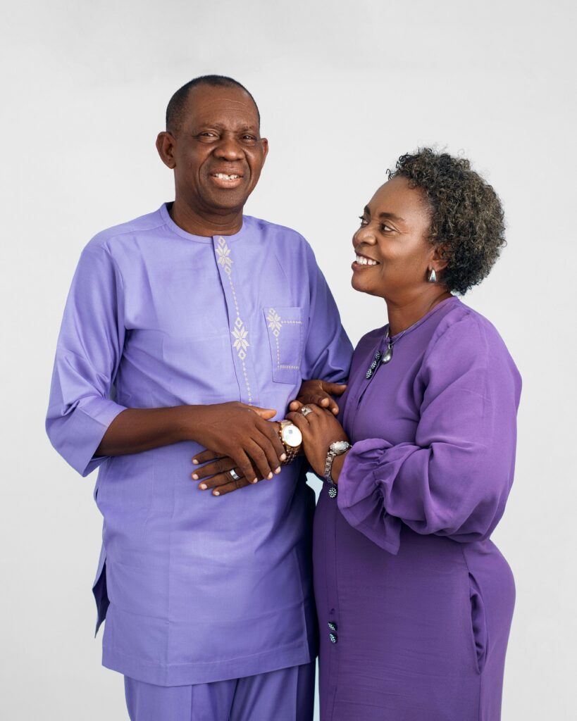 Elderly couple in matching purple outfits smiling joyfully at each other in a studio setting.